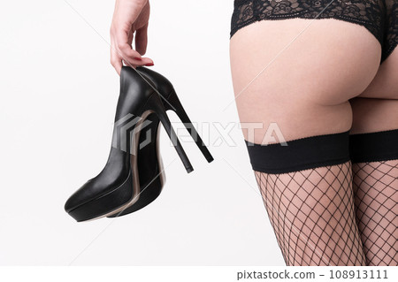 Rear view of woman holds pair of high heels in hand at waist level. Female in panties and stockings 108913111