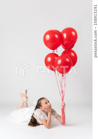 Thoughtful girl ten years old dressed in white dress holding lot of red balloons in hand, looking up 108913291
