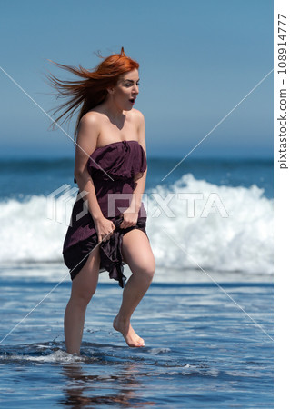 Happy and enthusiastic woman in long dress running through water of breaking ocean waves on beach 108914777