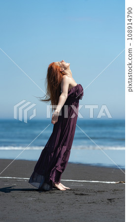 Happy woman in long dress stands on beach with head looking upward, eyes closed on background of sea 108914790
