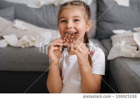Portrait of little girl eating chocolate at home. Background of scattered napkins. Chaos at home. Mess. Sweets 108925288