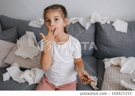 Adorable and beautiful little girl eats chocolate, but it melts in her hands, so the girl licks her fingers. Sweets. Chaos on background. Mess 108925355