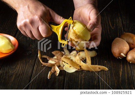 The chef peels raw potatoes in a hotel kitchen before preparing a national dish. Close-up of a cook hands with a potato peeler while working 108925921