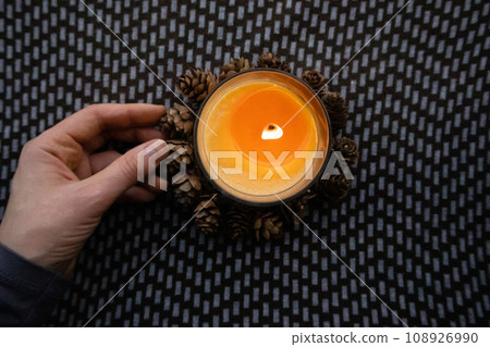 Yellow round candle in a woman's hand on a dark ornamental background flatly top view. Home decor details. Woman holding a scented aromatic burning candle among pine cones. Touch a fire. Warm light. 108926990