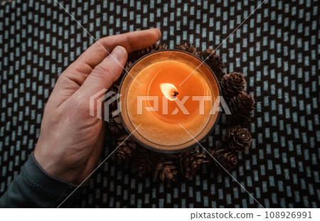 Yellow round candle in a woman's hand on a dark ornamental background flatly top view. Home decor details. Woman holding a scented aromatic burning candle among pine cones. Touch a fire. Warm light. Yellow round candle in a woman's hand on a dark ornamental background flatly top view. Home decor details. Woman holding a scented aromatic burning candle among pine cones. Touch a fire. Warm light. 108926991