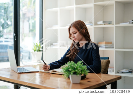 A beautiful caucasian female student is studying in college remotely. She is sitting at a table at home with a laptop and a notepad and concentrated is watching a video conference lesson 108928489