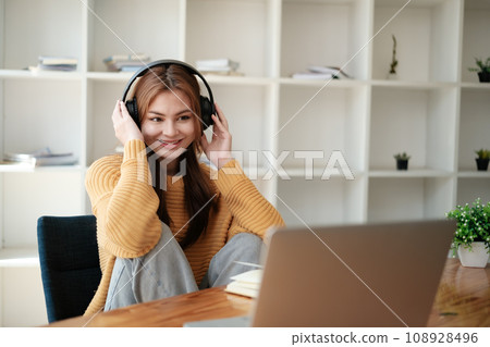 A beautiful caucasian female student is studying remotely. She is sitting at a table at home with a laptop and a notepad and concentrated is watching a video conference, smiling. 108928496