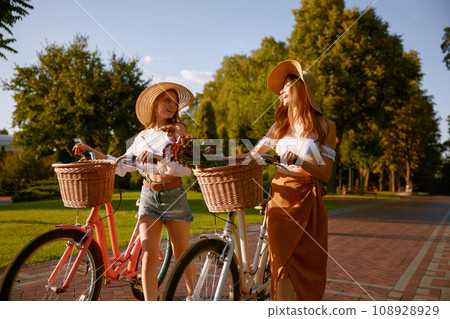 Two girl best friends nice conversation while walking with bicycle at park Two girl best friends nice conversation while walking with bicycle at park 108928929