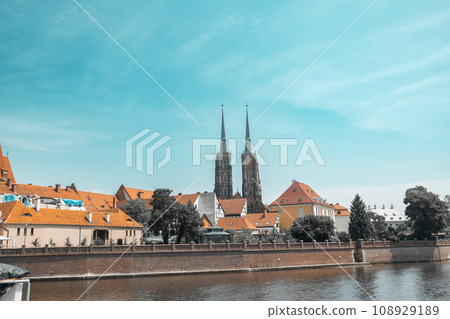 Street with cobblestone road with green trees, colorful buildings, summer cafe, Cathedral of St. John the Baptist church with two spires in old historical city centre, Ostrow Tumski, Wroclaw, Poland 108929189