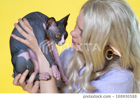 Young blonde cosplay elf in blue dress holding Sphinx kitten in hands, soothes it with her smile Young blonde cosplay elf in blue dress holding Sphinx kitten in hands, soothes it with her smile 108930388