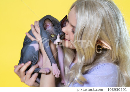 Cosplayer elf young female in blue dress holds Sphinx kitten in hands, hugs it and kisses gently Cosplayer elf young female in blue dress holds Sphinx kitten in hands, hugs it and kisses gently 108930391