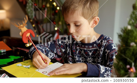 Closeup of little boy writing a letter to Santa in a living room decorated for Christmas 108930550