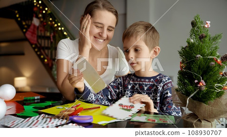 Portrait of happy smiling mother looking at her son making Christmas postcards and writing a letter to Santa 108930707