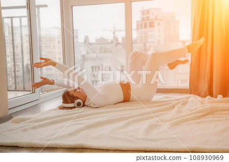 Side view portrait of relaxed woman listening to music with headphones lying on carpet at home. She is dressed in a white tracksuit. Side view portrait of relaxed woman listening to music with headphones lying on carpet at home. She is dressed in a white tracksuit. 108930969