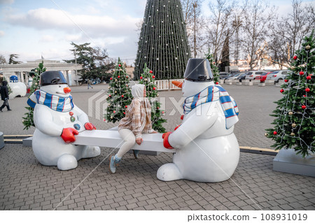 Woman Christmas Square. Close-up of a winter white toy snowman With trees decorated with Christmas tinsel in the background Woman Christmas Square. Close-up of a winter white toy snowman With trees decorated with Christmas tinsel in the background 108931019