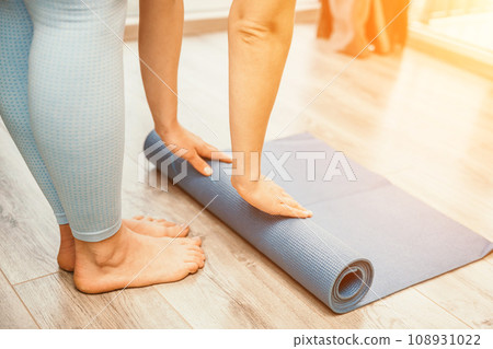 Woman hands rolled up yoga mat on gym floor in yoga fitness training room. Home workout woman close up hands rolling foam yoga gym mat. Woman barefoot home workout sportive healthy lifestyle concept 108931022