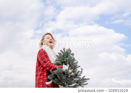 Blond woman holding Christmas tree by the sea. Christmas portrait of a happy woman walking along the beach and holding a Christmas tree in her hands. Dressed in a red coat, white suit. 108931029