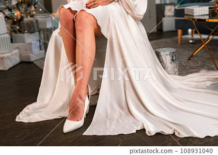 Women legs christmas tree. Cropped photo of beautiful elegant female legs in white high-heeled shoes and a white dress, a woman sits against the background of a Christmas tree with gifts. Women legs christmas tree. Cropped photo of beautiful elegant female legs in white high-heeled shoes and a white dress, a woman sits against the background of a Christmas tree with gifts. 108931040