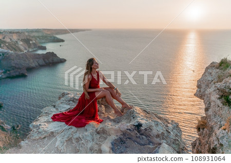 Woman sunset sea red dress, side view a happy beautiful sensual woman in a red long dress posing on a rock high above the sea on sunset. Woman sunset sea red dress, side view a happy beautiful sensual woman in a red long dress posing on a rock high above the sea on sunset. 108931064