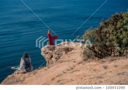 A woman in a red flying dress fluttering in the wind, against the backdrop of the sea. 108931090