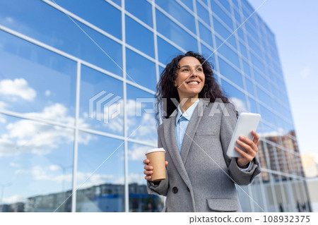 Successful Hispanic businesswoman walks to the office, holds a coffee and a phone in her hands, smiles and solves cases on the way to work, stands on the street of the office. 108932375
