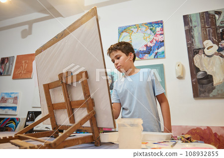 Teenage boy concentrated on drawing on canvas, standing by wooden easel at art class, against painted pictures backdrop 108932855