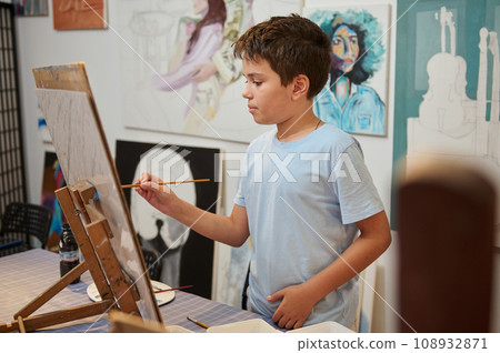 Adorable Caucasian child boy holding paintbrush, drawing on canvas in creative workshop or elementary age school 108932871