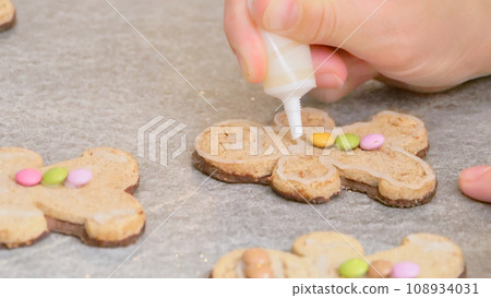 woman's hands decorating a gingerbread man-shaped Christmas cake with white icing and colored caramels, close-up 108934031