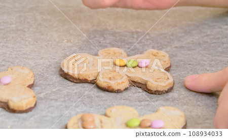 woman's hands decorating a gingerbread man-shaped Christmas cake with white icing and colored caramels, close-up 108934033