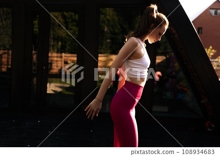 Side view of redhead woman rotating shoulders doing warm up exercise during fitness training, standing on background of house on sunny summer morning. Female training in open air warms joints. Side view of redhead woman rotating shoulders doing warm up exercise during fitness training, standing on background of house on sunny summer morning. Female training in open air warms joints. 108934683