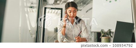 Woman office manager talking phone with client and making notes while sitting the desk in office Woman office manager talking phone with client and making notes while sitting the desk in office 108934792