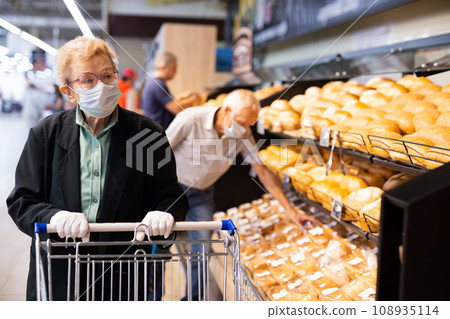 Older woman with glasses chooses buns and bread in supermarket bakery Older woman with glasses chooses buns and bread in supermarket bakery 108935114