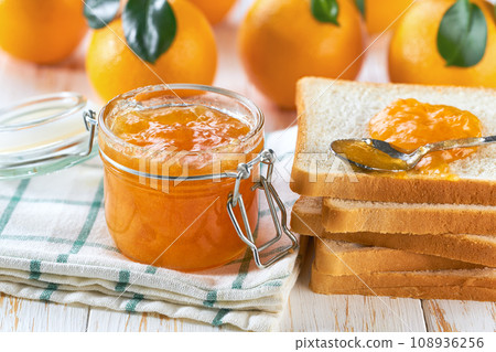 Homemade jam. A glass jar of orange jam on a white wooden table. Homemade orange marmalade and fresh oranges and green leaf on the kitchen table. Homemade jam. A glass jar of orange jam on a white wooden table. Homemade orange marmalade and fresh oranges and green leaf on the kitchen table. 108936256
