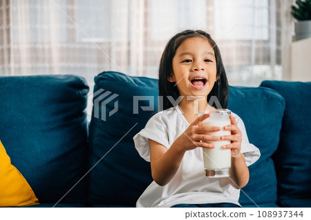 A delightful portrait of an Asian child holding a glass of milk smiling with joy while seated on the sofa. This photo emphasizes the importance of nurturing children with calcium-rich milk. A delightful portrait of an Asian child holding a glass of milk smiling with joy while seated on the sofa. This photo emphasizes the importance of nurturing children with calcium-rich milk. 108937344