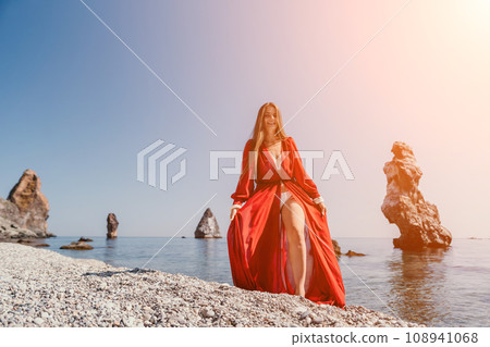 Woman travel sea. Happy tourist in red dress enjoy taking picture outdoors for memories. Woman traveler posing on the rock at sea bay surrounded by volcanic mountains, sharing travel adventure journey Woman travel sea. Happy tourist in red dress enjoy taking picture outdoors for memories. Woman traveler posing on the rock at sea bay surrounded by volcanic mountains, sharing travel adventure journey 108941068