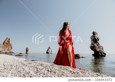 Woman travel sea. Happy tourist in red dress enjoy taking picture outdoors for memories. Woman traveler posing on the rock at sea bay surrounded by volcanic mountains, sharing travel adventure journey Woman travel sea. Happy tourist in red dress enjoy taking picture outdoors for memories. Woman traveler posing on the rock at sea bay surrounded by volcanic mountains, sharing travel adventure journey 108941072