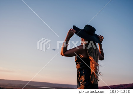 Woman lavender field. Happy carefree woman in black dress and hat with large brim walking in a lavender field during sunset. Perfect for inspirational and warm concepts in travel and wanderlust. Woman lavender field. Happy carefree woman in black dress and hat with large brim walking in a lavender field during sunset. Perfect for inspirational and warm concepts in travel and wanderlust. 108941238
