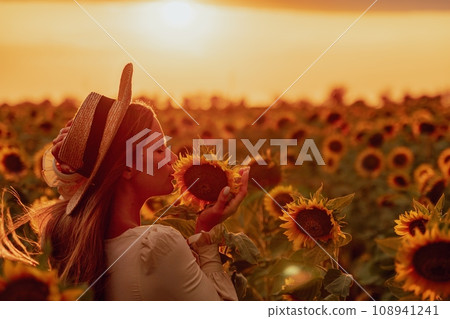 Woman in Sunflower Field: Happy girl in a straw hat posing in a vast field of sunflowers at sunset, enjoy taking picture outdoors for memories. Summer time. Woman in Sunflower Field: Happy girl in a straw hat posing in a vast field of sunflowers at sunset, enjoy taking picture outdoors for memories. Summer time. 108941241