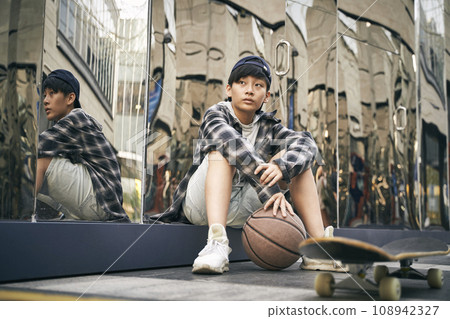 teenage asian kid taking a break sitting on ground with basketball and skateboard teenage asian kid taking a break sitting on ground with basketball and skateboard 108942327