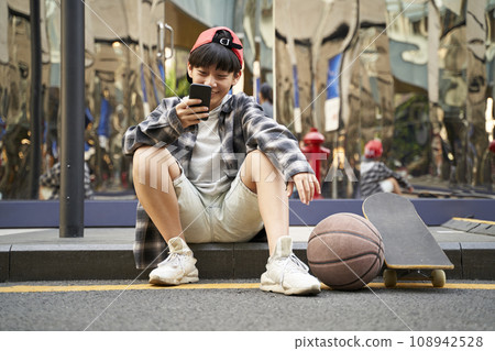 teenage asian kid with basketball and skateboard sitting on curb of street looking at cellphone teenage asian kid with basketball and skateboard sitting on curb of street looking at cellphone 108942528