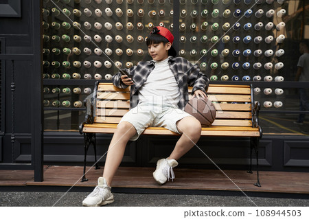 fifteen-year-old teenage asian boy sitting on bench looking at cellphone fifteen-year-old teenage asian boy sitting on bench looking at cellphone 108944503