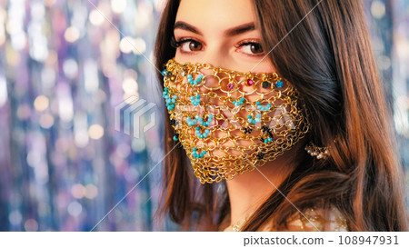 Pandemic fashion. DIY accessory. Closeup of mysterious woman with night makeup in matching glamour gold chain face mask on blur blue sparkling tinsel background. Pandemic fashion. DIY accessory. Closeup of mysterious woman with night makeup in matching glamour gold chain face mask on blur blue sparkling tinsel background. 108947931