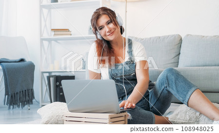Enjoying music. Romantic woman. Home relax. Smiling middle-aged lady in headphones sitting floor scrolling laptop in light room interior. Enjoying music. Romantic woman. Home relax. Smiling middle-aged lady in headphones sitting floor scrolling laptop in light room interior. 108948025