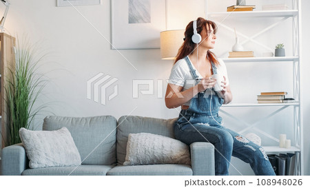 Enjoying music. Romantic woman. Coffee time. Middle-aged lady in headphones sitting sofa with cup of beverage light room interior copy space. 108948026