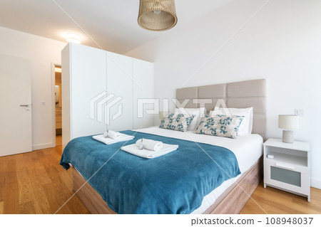 Double bed in bright bedroom room interior furnishing furniture. With blue blanket and towels in a roll of fragrant grass and toilet in background. Double bed in bright bedroom room interior furnishing furniture. With blue blanket and towels in a roll of fragrant grass and toilet in background. 108948037