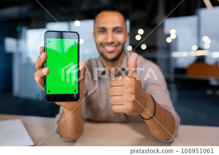 Happy man sitting in office holding phone with green screen and showing thumb up, phone in foreground. 108951061