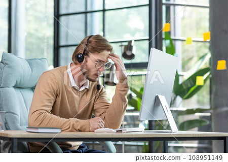 Tired worker in headset holding hand to head, sitting at table, suffering from headache, tired from work wants to sleep. Tired worker in headset holding hand to head, sitting at table, suffering from headache, tired from work wants to sleep. 108951149