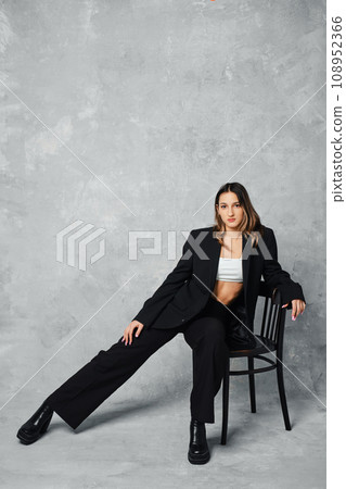 Stylish young woman in black pantsuit and ankle boots sitting on chair put one leg aside 108952366