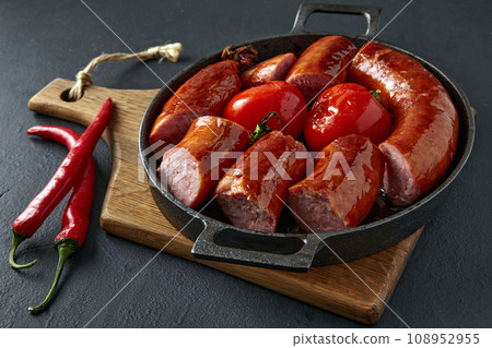 Fried pork sausage cut into pieces and grilled tomatoes on a black cast iron frying pan with cutlery and hot peppers on a black stone table Fried pork sausage cut into pieces and grilled tomatoes on a black cast iron frying pan with cutlery and hot peppers on a black stone table 108952955