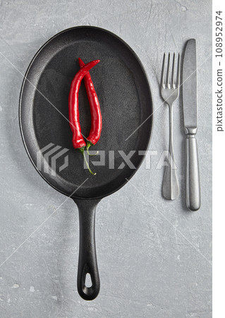 Two hot peppers on a black cast iron frying pan and cutlery on the background of a stone table. Top view with copy space 108952974